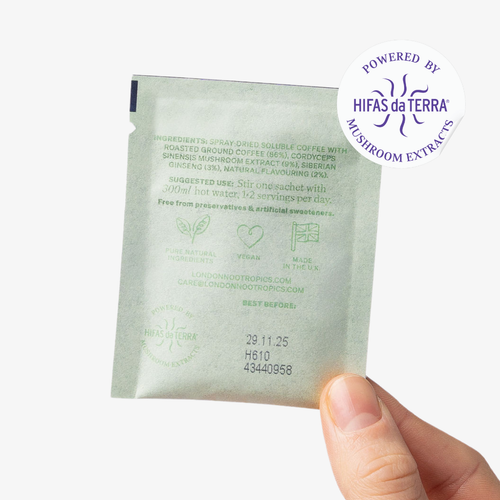 Hand holding a single-serve green sachet of London Nootropics Mojo coffee. The images shows the backside of the sachet which features an ingredient list and the best before date. The image shows a sticker icon at the top right corner of the sachet that reads: Powered by Hifas da Terra Mushroom Extracts.