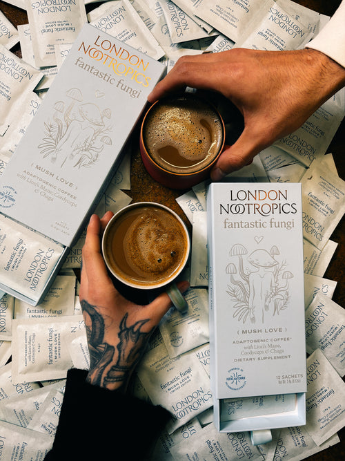 Overhead image of a surface covered in single-serve Mush Love sachets. Two coffee mugs full of London Nootropics coffee sit in the middle, held by hands coming from the opposite ends of the image.