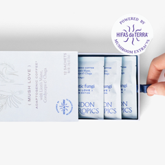 Slightly opened box of Mush Love Lion's Manne, Cordyceps, and Chaga adaptogenic coffee showing three single serve sachets. A hand pull the box open. In the top right corner is a sticker that reads: Powered by Hifas da Terra mushroom extracts.