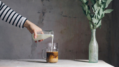 Arm with striped white and black sleeve reaches towards the centre of the image to pour a glass of milk into a mug of London Nootropics mushroom coffee.