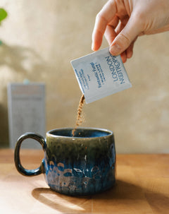 Image of hand pouring mushroom coffee from a blue Mush Love blend single-serve sachet into a ceramic mug. The image is set indoors with warm lighting.
