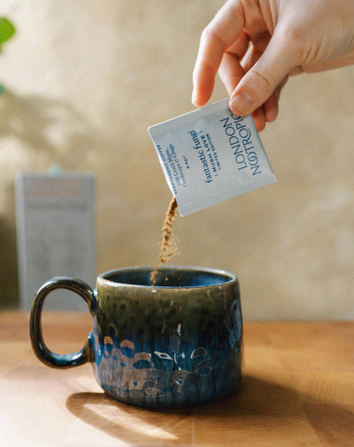 Image of hand pouring mushroom coffee from a blue Mush Love blend single-serve sachet into a ceramic mug. The image is set indoors with warm lighting.
