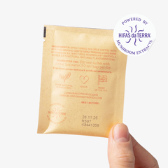 Hand holding a single-serve tan coloured sachet of London Nootropics Flow coffee. The image shows a sticker icon at the top right corner of the sachet that reads: Powered by Hifas da Terra Mushroom Extracts. The backside of the sachets displays an ingredient list and best before date.