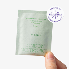 Hand holding a single-serve green sachet of London Nootropics Mojo adaptogenic coffee. The image shows a sticker icon at the top right corner of the sachet that reads: Powered by Hifas da Terra Mushroom Extracts.
