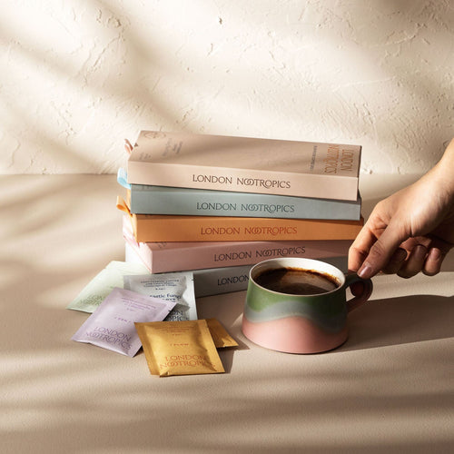 Stack of pastel coloured london nootropics coffee boxes on a minimalistic white background with gentle shadows casted from an angle. Next to the stack of boxes is an assortment of London Nootropics adaptogenic coffee blends and a mug full of mushroom coffee.